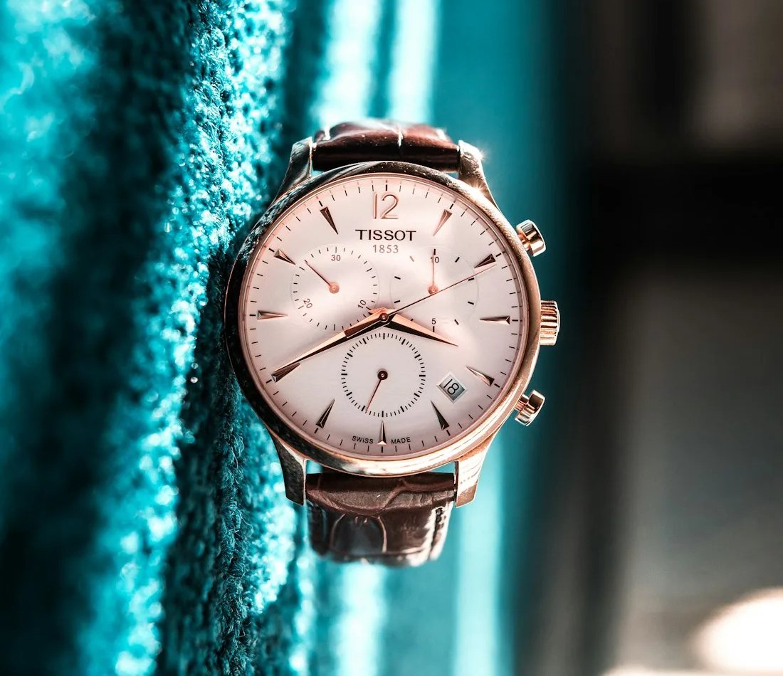 Rose-gold chronograph with a white dial and brown leather strap resting on teal fabric